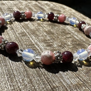 Pode incluir: Uma pulseira de contas rosa, bordô e opalescentes. A pulseira apresenta contas redondas em tons de rosa e bordô, intercaladas com contas opalescentes e espaçadores prateados. A pulseira é exibida em uma superfície de madeira.
