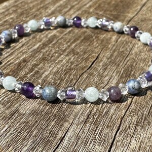 May include: A beaded bracelet with amethyst, aquamarine, and clear quartz beads. The bracelet is on a wooden surface.