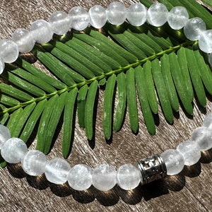 May include: A white crystal beaded bracelet with a silver charm. The bracelet is on a wooden surface with a green leaf.