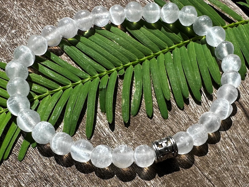May include: A white crystal beaded bracelet with a silver charm. The bracelet is on a wooden surface with a green leaf.