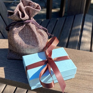 May include: A light blue gift box with a brown satin ribbon tied in a bow. The box is sitting on a wooden surface next to a brown burlap bag.