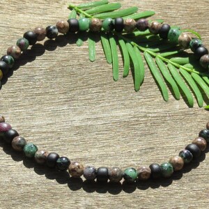 May include: A beaded bracelet featuring a mix of matte black, green, brown, and reddish-purple beads. The bracelet is strung together, creating a circular shape. The background is a wooden surface with green leaves.