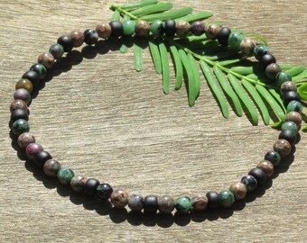 May include: A beaded bracelet featuring a mix of matte black, green, brown, and reddish-purple beads. The bracelet is strung together, creating a circular shape. The background is a wooden surface with green leaves.