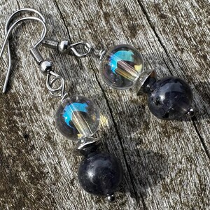 Peut inclure: Une paire de boucles d'oreilles pendantes avec des crochets argentés. Chaque boucle d'oreille comporte une perle de verre transparente et irisée avec une teinte bleue, une entretoise argentée et une perle sphérique gris foncé. Sur une surface en bois.