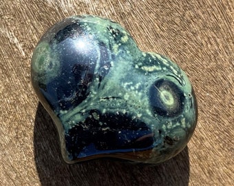 Puede incluir: Una piedra verde y negra en forma de corazón con patrones en espiral y motas blancas. La piedra tiene una superficie lisa y pulida.
