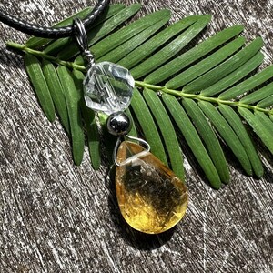 May include: A pendant necklace featuring a clear, faceted crystal, a small silver bead, and a teardrop-shaped amber-colored stone. The pendant hangs from a black cord. The necklace rests on a green leaf against a textured, wooden surface.