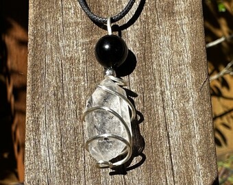 May include: A clear quartz crystal wrapped in silver wire, hanging from a black cord with a black bead. The pendant is displayed against a wooden background.