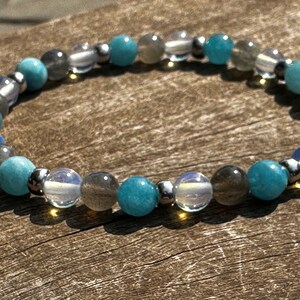 Pode incluir: Um bracelete feito com contas azuis, cinzas e brancas. O bracelete é feito com pedras naturais e detalhes metálicos.