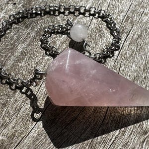 Può includere: Un pendolo di quarzo rosa rosa con una catena d'argento e una perla di quarzo trasparente. Il pendolo è su una superficie di legno.