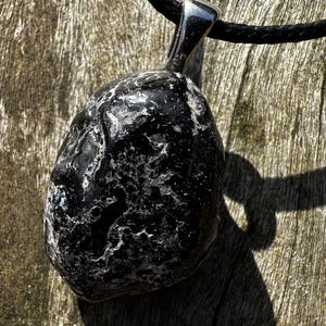 Può includere: Un ciondolo in pietra nera di forma irregolare con una superficie lucida e strutturata caratterizzata da inclusioni minerali bianche. Il ciondolo è fissato a un gancio in metallo color argento e pende da un cordino nero, creando un contrasto.