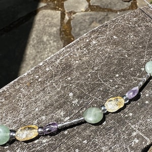 May include: A necklace featuring a string of polished gemstones in shades of green, yellow, and purple. The stones are interspersed with silver-colored metal beads and tubes. The necklace is displayed on a weathered wooden surface.