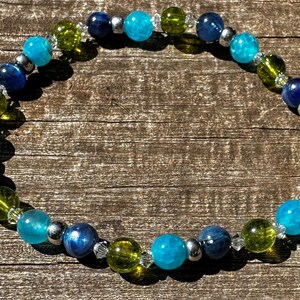 May include: A beaded bracelet featuring a mix of blue, green, and silver beads. The bracelet includes turquoise, dark blue, and olive green beads, interspersed with silver-colored spacers and clear crystal accents. The beads are strung on an elastic band.
