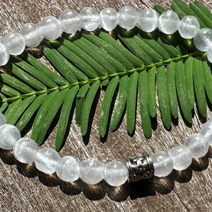 May include: A white crystal beaded bracelet with a silver charm. The bracelet is on a wooden surface with a green leaf.