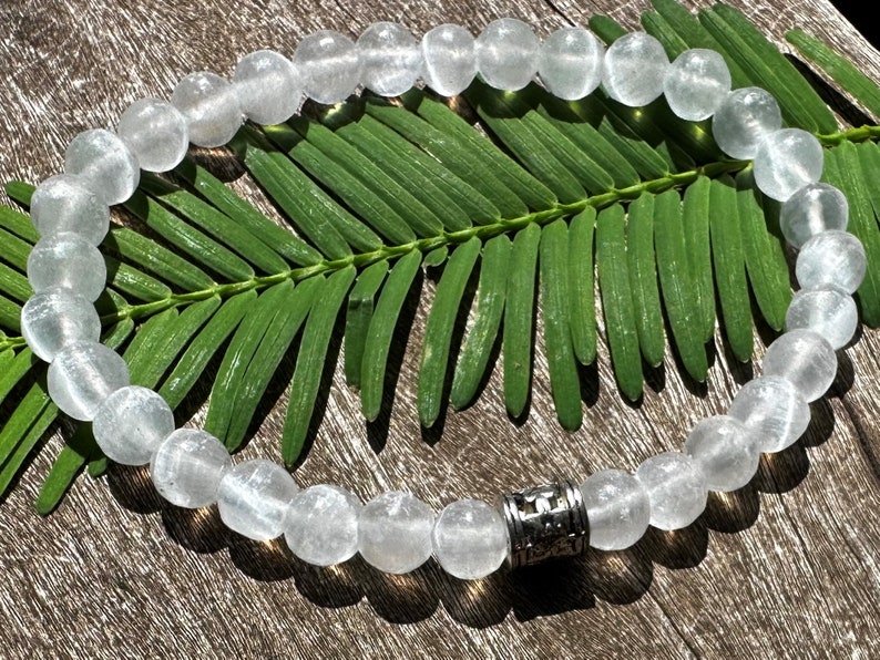 May include: A white crystal beaded bracelet with a silver charm. The bracelet is on a wooden surface with a green leaf.