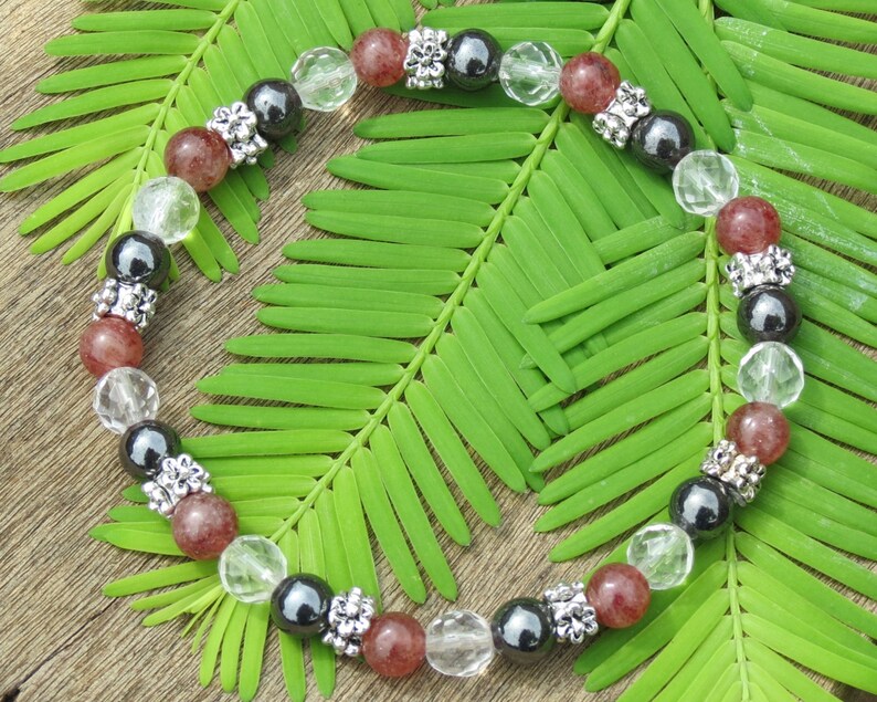 K&ouml;nnte beinhalten: Ein kreisf&ouml;rmiges Armband mit Perlen in transparenten, schwarzen und rotbraunen Farbt&ouml;nen, durchsetzt mit silberfarbenen Zierelementen. Das Armband liegt auf einem Bett aus gr&uuml;nen Bl&auml;ttern und bildet einen nat&uuml;rlichen Hintergrund.