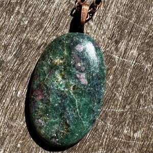 May include: A green and pink oval-shaped gemstone pendant on a copper chain. The pendant has a mottled green and pink color with a smooth, polished surface.