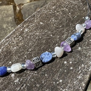 May include: A beaded necklace featuring a variety of gemstones. The necklace includes blue, purple, and white stones, along with silver-colored square beads. The stones are strung on a black cord, creating a unique and colorful accessory.