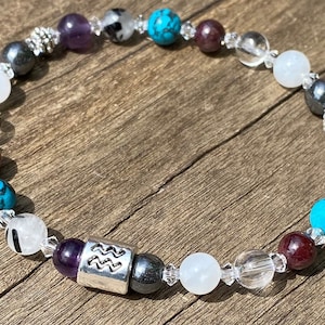 May include: A beaded bracelet with a silver charm featuring the astrological symbol for Aquarius. The bracelet features clear, white, purple, red, and turquoise beads.