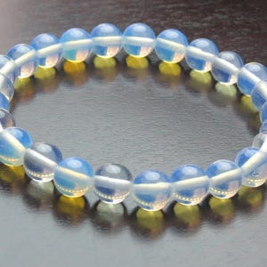 Puede incluir: Un brazalete hecho de cuentas de ópalo iridiscente. Las cuentas son de color azul claro con un brillo iridiscente blanco.