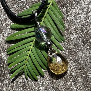 May include: A pendant necklace with a black cord. The pendant features a clear, round bead with iridescent accents, a small silver bead, and a teardrop-shaped bead with golden inclusions. The necklace rests on a green leaf against a wood-grain background.