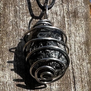 Op de afbeelding: Een zwarte stenen hanger, omwikkeld met zilverkleurig draad, hangt aan een zwart koord. De draad is in een spiraal om de steen gewikkeld. De hanger staat tegen een verweerde houten achtergrond.