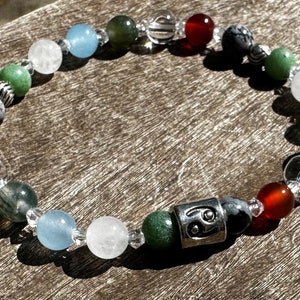 May include: A beaded bracelet with a silver charm featuring the astrological symbol for Aquarius. The bracelet features clear, red, green, and blue beads.
