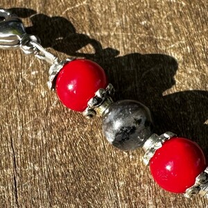 May include: A silver charm with a red and gray bead design. The charm has a lobster clasp and is attached to a silver chain.