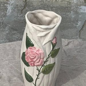 Puede incluir: Un jarrón de cerámica color crema con una superficie texturizada y un diseño floral. El jarrón presenta una rosa rosa con hojas verdes y un capullo en un tallo delgado. El jarrón mide aproximadamente 20 cm de alto.