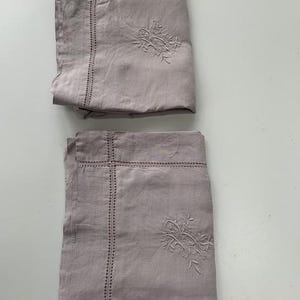 May include: Two folded, dusty rose-colored linen pillowcases. Each pillowcase features an embroidered floral design and a decorative stitched border. The pillowcases are made of a soft, natural fabric.