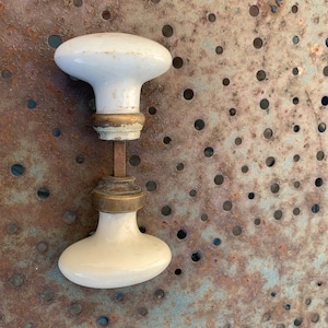 Puede incluir: Un par de pomos de puerta antiguos de porcelana blanca con detalles de latón. Los pomos ovalados están conectados por una varilla metálica. El fondo es una chapa metálica oxidada y perforada.