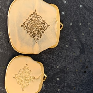 Peut inclure: Deux poudriers dorés ornés d'un motif de filigrane décoratif au centre. Les poudriers ont un bord festonné et un petit fermoir doré. Le fond est une surface sombre et texturée avec de petits points argentés.