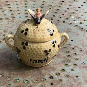 Può includere: Un vasetto di miele in ceramica con un motivo a nido d'ape strutturato e dettagli neri. Il coperchio è sormontato da una figura di ape. La parola "meli" è stampata sulla parte anteriore del vasetto. Il vasetto ha due piccole maniglie.