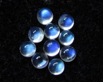 Puede incluir: Un grupo de 10 cabujones redondos de piedra lunar iridiscente con un brillo azul. Las piedras están dispuestas en un grupo suelto sobre un fondo negro.