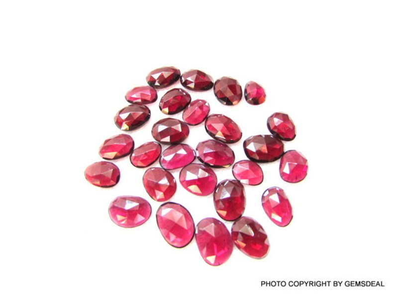 May include: A collection of 20 oval-shaped, faceted, red gemstones. The gemstones are scattered on a white surface.