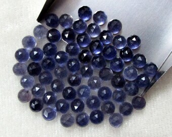 May include: A collection of faceted, round, deep blue gemstones. The gems are scattered across a white surface and a silver tray. The gems have a polished, reflective surface, and are likely used for jewelry making or collecting.