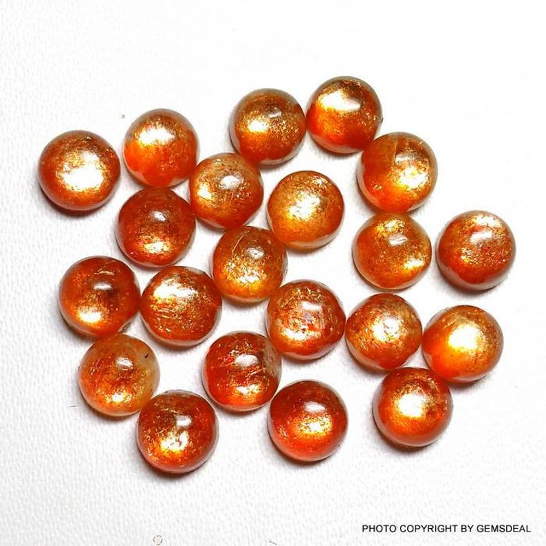 May include: A collection of 20 round, orange sunstone cabochons. The stones have a metallic sheen and are arranged in a random pattern.