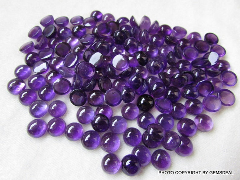 May include: A large group of round, purple amethyst cabochons. The cabochons are smooth and polished, with a slight sheen.