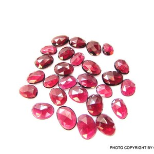 May include: A collection of 20 oval-shaped, faceted, red gemstones. The gemstones are scattered on a white surface.
