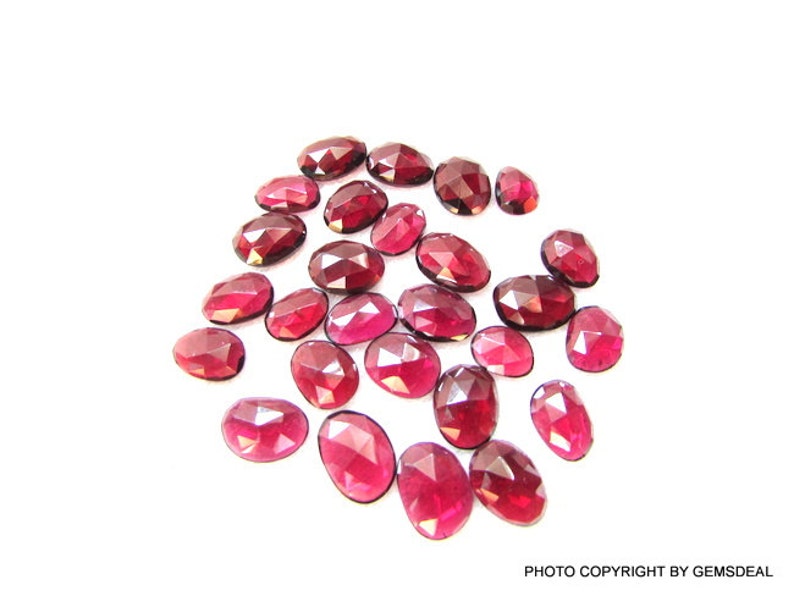 May include: A collection of 20 oval-shaped, faceted, red gemstones. The gemstones are scattered on a white surface.