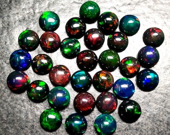 May include: A collection of 25 round, black opal cabochons with iridescent green, blue, and red flashes. The opals are arranged in a random pattern on a gray surface.