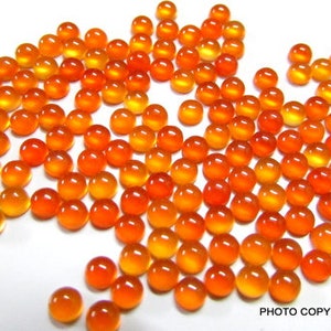 May include: A large group of orange and yellow cabochon gemstones, likely carnelian, scattered on a white surface. The gemstones are round and smooth, with a polished finish.