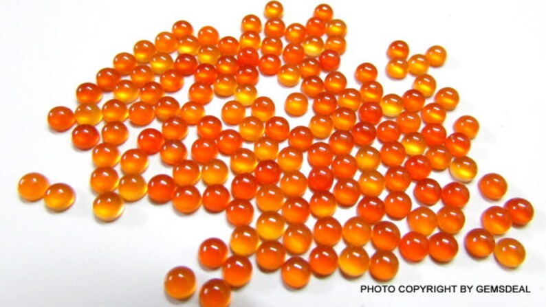 May include: A large group of orange and yellow cabochon gemstones, likely carnelian, scattered on a white surface. The gemstones are round and smooth, with a polished finish.