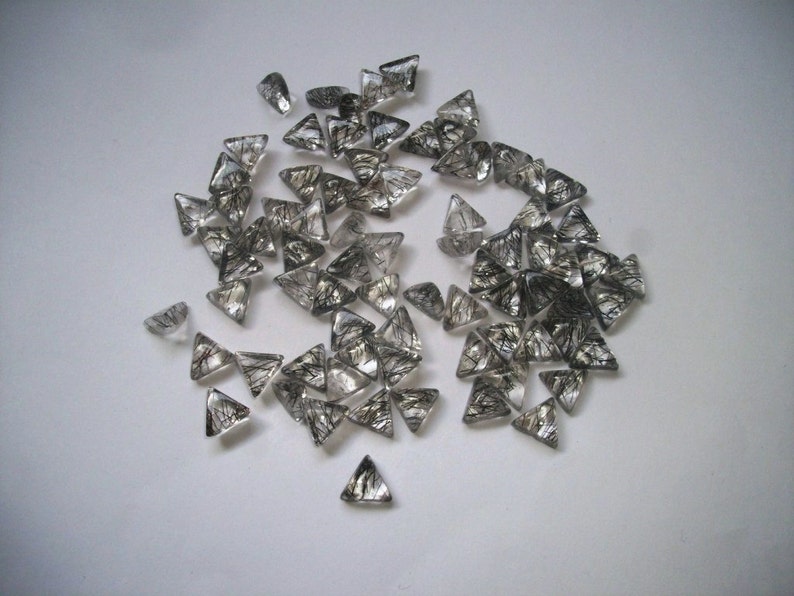 May include: A collection of clear quartz crystal points with black inclusions, also known as rutilated quartz, in a triangular shape. The crystals are scattered on a white surface.