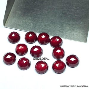 May include: A collection of faceted, deep red gemstones, possibly rubies, arranged on a white surface. The gems have a rounded shape with multiple facets, reflecting light. A metal tray is in the background. The text "GEMSDEL" is visible.