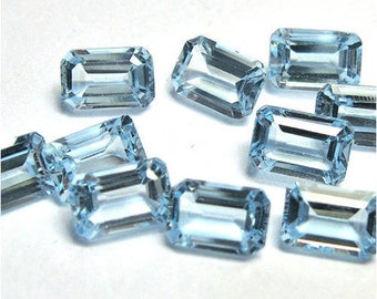 May include: A group of ten light blue emerald cut gemstones. The gemstones are faceted and have a clear, glassy appearance.