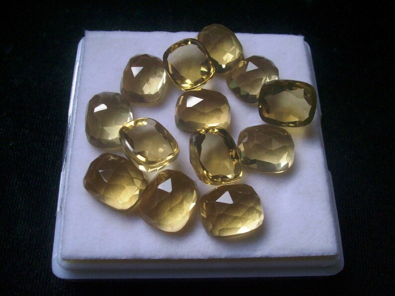May include: A collection of 12 faceted, yellow gemstones displayed on a white background. The gemstones are various sizes and shapes, with some being more rounded than others.