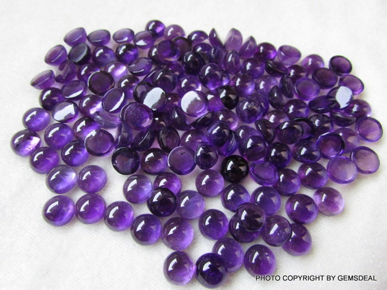 May include: A large pile of round, purple amethyst cabochons. The cabochons are smooth and polished, with a deep purple color.