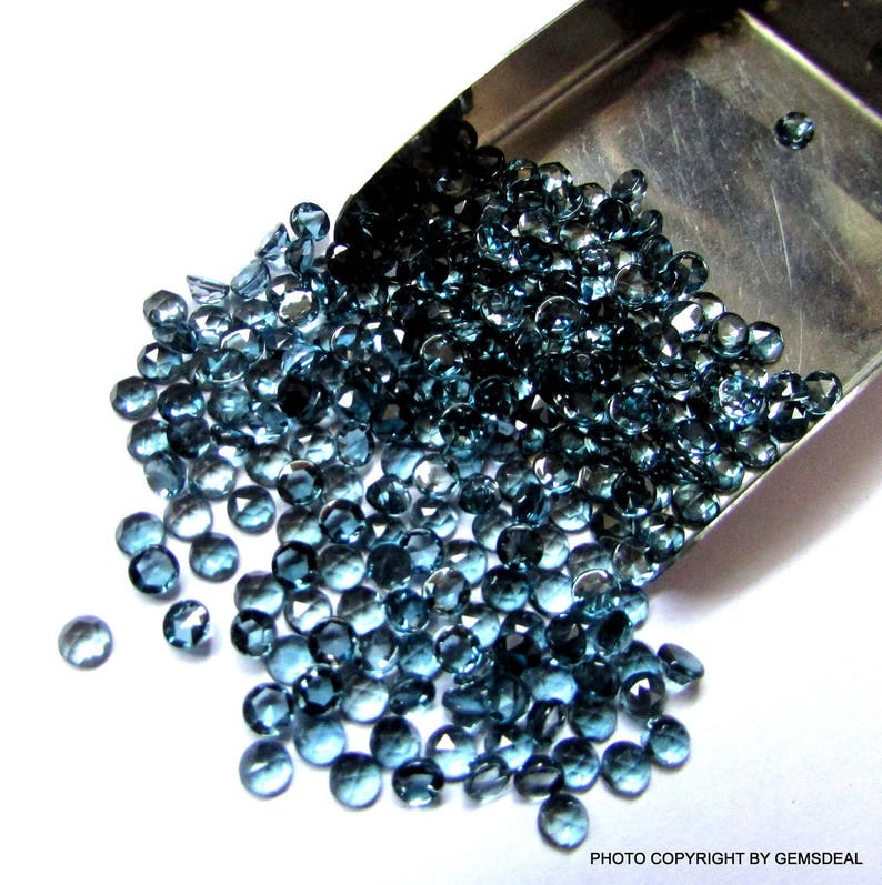 K&ouml;nnte beinhalten: Eine Schaufel gef&uuml;llt mit losen blauen Topas-Edelsteinen. Die Edelsteine sind rund und facettiert und liegen auf einer wei&szlig;en Oberfl&auml;che.
