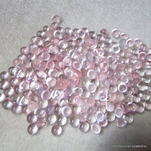May include: A large pile of pale pink and clear round cabochon gemstones. The gemstones are smooth and polished, and they have a translucent appearance.