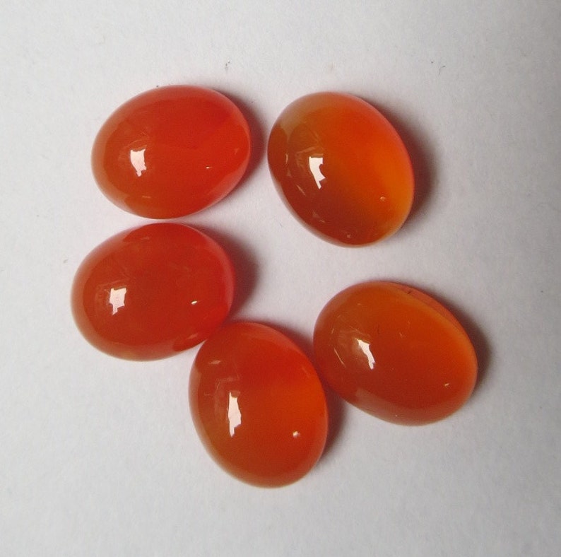 May include: Five oval-shaped carnelian gemstones, each with a smooth, polished surface and a vibrant orange color.
